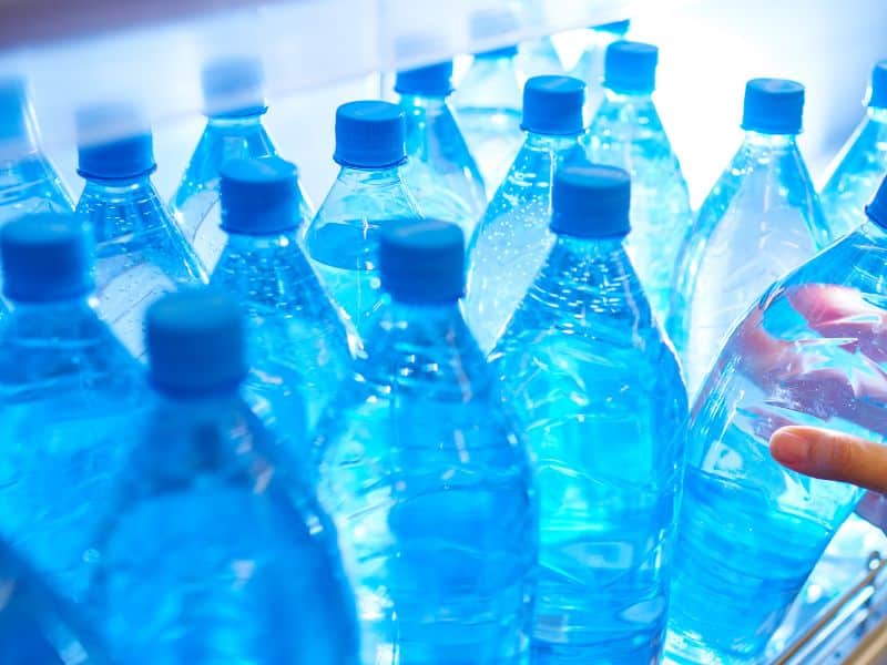 A selection of blue plastic water bottles with caps, closely packed together with a hand reaching for one, illuminated by a cool blue light that gives the scene a fresh and clean feeling.