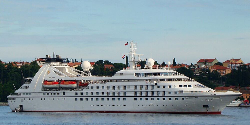 The luxury cruise ship Seabourn Spirit gliding through calm waters near a coastal town. The vessel is predominantly white with sleek, streamlined contours and includes multiple decks visible from this side angle. Prominent features are lifeboats housed mid-ship and a satellite dome above. The backdrop shows a serene waterfront with scattered red-roofed houses, highlighting a picturesque, suburban coastline setting.