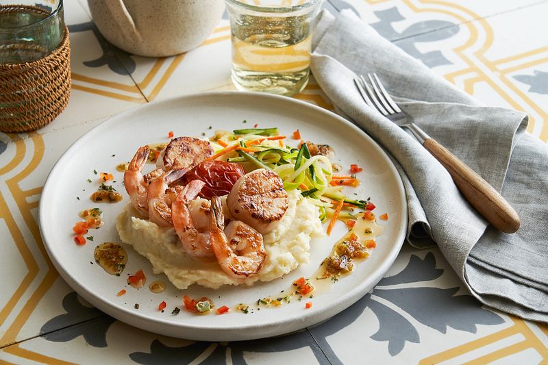 A gourmet meal from the Main Dining Room Menu of a Princess Cruise ship, artfully plated with succulent seared scallops and shrimp atop creamy mashed potatoes, garnished with julienne vegetables, served on a stylish white plate with a patterned tablecloth setting.
