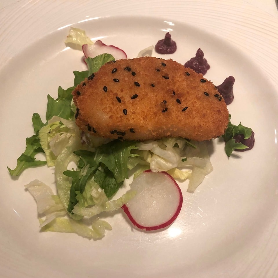 A vegetarian dinner served in the main dining room on the P&O Britannia. The dish features a breaded and fried patty with black sesame seeds, placed on a bed of mixed greens and thinly sliced radishes, garnished with small dollops of purple sauce. The presentation is elegant and appetizing.