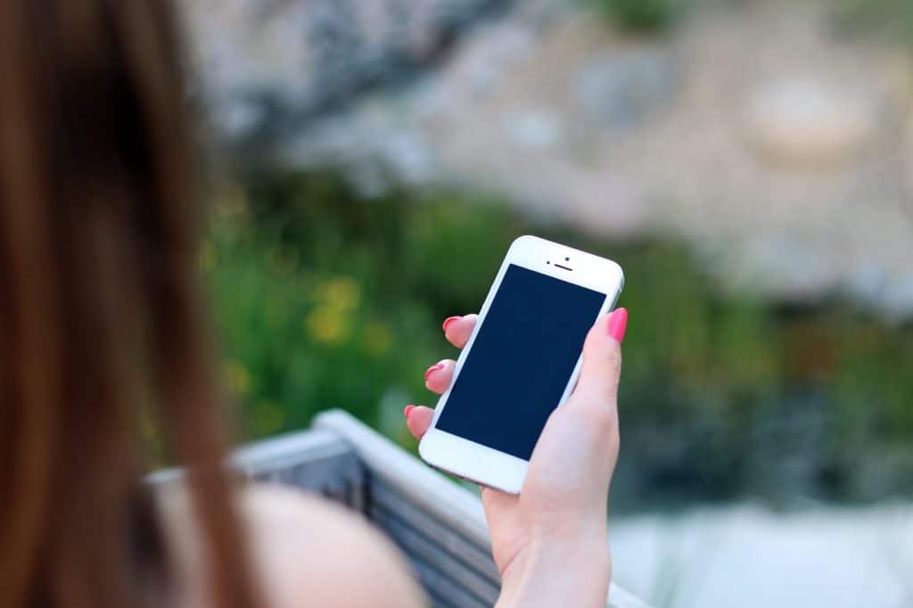 A woman with pink-painted nails is holding a white smartphone with a blank screen, as she appears to be outdoors. The background is blurred, showing greenery and a natural setting, suggesting she might be enjoying some time outside while using her phone. The focus is on the phone and her hand, emphasizing the casual, everyday use of technology.