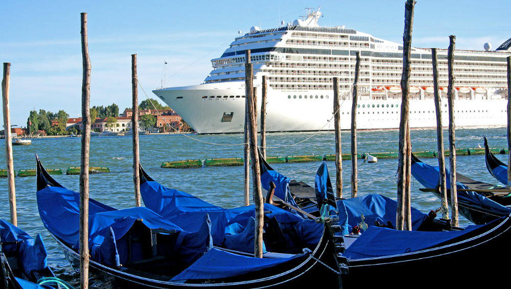 This image shows a large white cruise ship passing through a waterway in Venice, Italy, juxtaposed with traditional Venetian gondolas docked in the foreground. The gondolas, covered with blue tarps, contrast sharply with the imposing size of the modern ship, highlighting the tension between traditional culture and modern tourism in Venice.