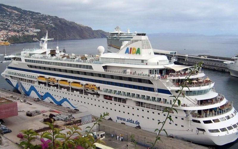 The AIDAvita cruise ship docked at port, with its distinctive eye-catching blue wave design along the hull, as passengers gather on the decks against a backdrop of a mountainous coastline.