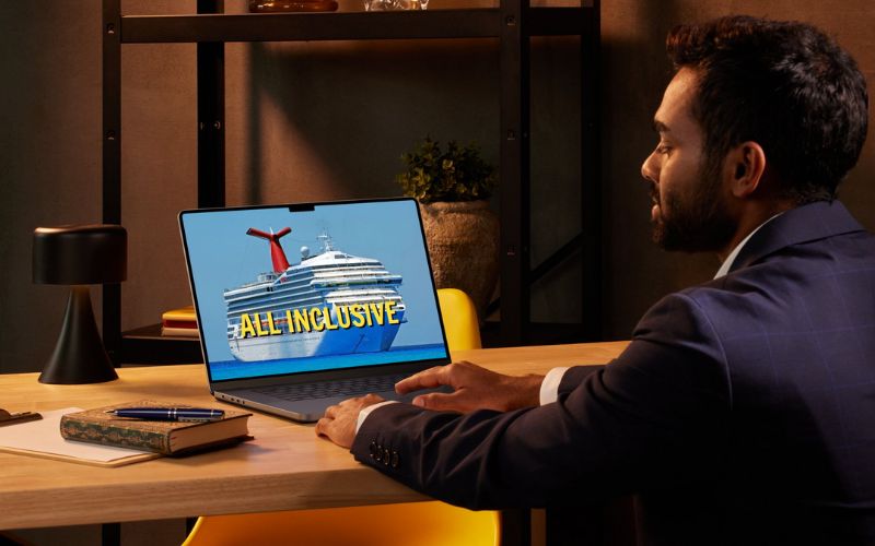 A man in a business suit sits at a desk, looking at his laptop screen which displays an image of a cruise ship with the words 'ALL INCLUSIVE' overlaid. The office setting includes a modern desk, a black lamp, and shelving units in the background.