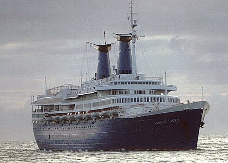 The ocean liner Achille Lauro, captured from a side angle as it navigates through the sea. The ship is characterized by its deep navy-blue hull and white superstructure, topped with two distinct blue funnels. The sea is moderately rough, emphasizing the vessel's maritime journey under a cloudy sky.