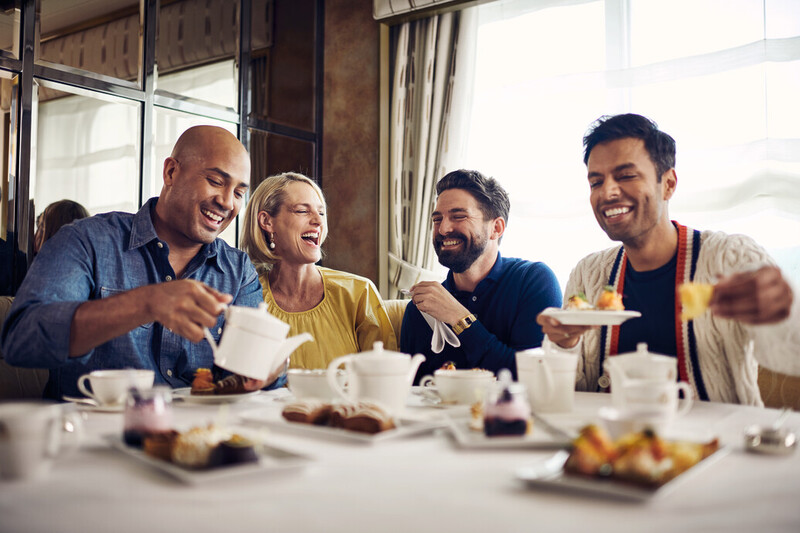 Four people enjoying a lively afternoon tea on a Cunard cruise ship. They're seated around a table with a white linen tablecloth, indulging in a variety of pastries and tea. Laughter and conversation are evident, as they share a cheerful moment, with one individual pouring tea and another offering a plate of food, encapsulating the joy of cruise dining experiences.