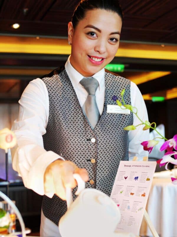 A smiling server in a patterned vest and tie pouring tea during afternoon tea service on a P&O cruise ship. The table is adorned with a menu and purple orchid decorations, adding a touch of elegance to the setting.