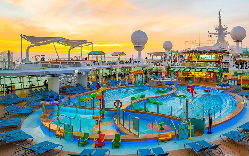 The image shows a colorful pool deck on a cruise ship during sunset. The area includes various pools, water slides, and play features surrounded by blue lounge chairs. Vibrant structures and shaded areas provide a lively and fun atmosphere, with the ocean visible in the background.