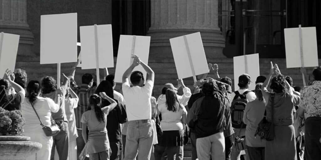 This is an image showing a group of people protesting, with their backs turned toward the camera and holding blank signs in front of a building with large columns. The black-and-white composition emphasizes the collective action, likely related to a social or political cause. If you'd like a specific focus for the alt text (e.g., "anti-tourism protests"), let me know!