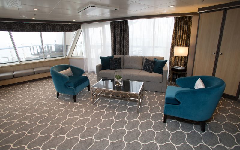 Elegant Aqua Theater Suite with Balcony on Harmony of the Seas, featuring a comfortable gray sofa, teal armchairs, and a patterned carpet, with natural light streaming through the large windows.