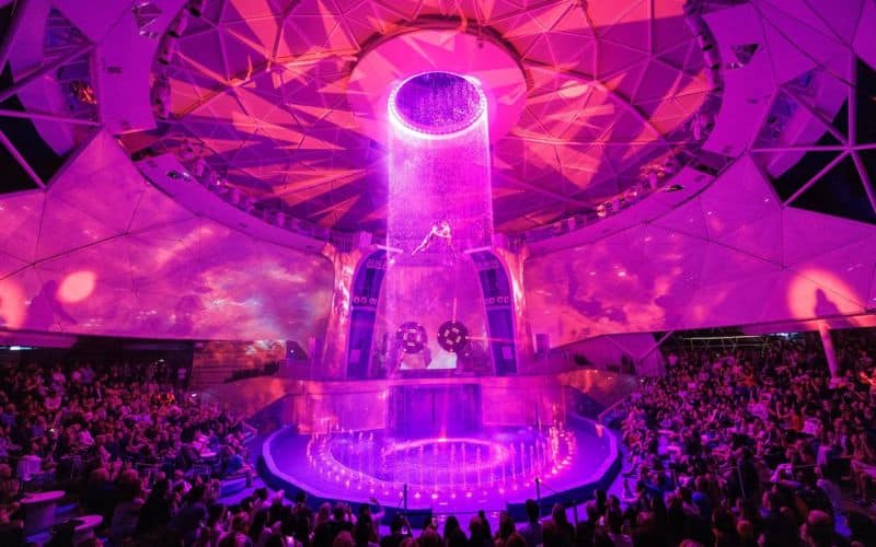 Spectacular performance at the AquaDome on Royal Caribbean's 'Star of the Seas.' The dome-shaped venue is bathed in vibrant purple lighting, featuring a circular water stage where an acrobat performs mid-air, surrounded by cascading water curtains. The audience, filling the surrounding seats, watches in awe, creating an immersive and theatrical atmosphere.