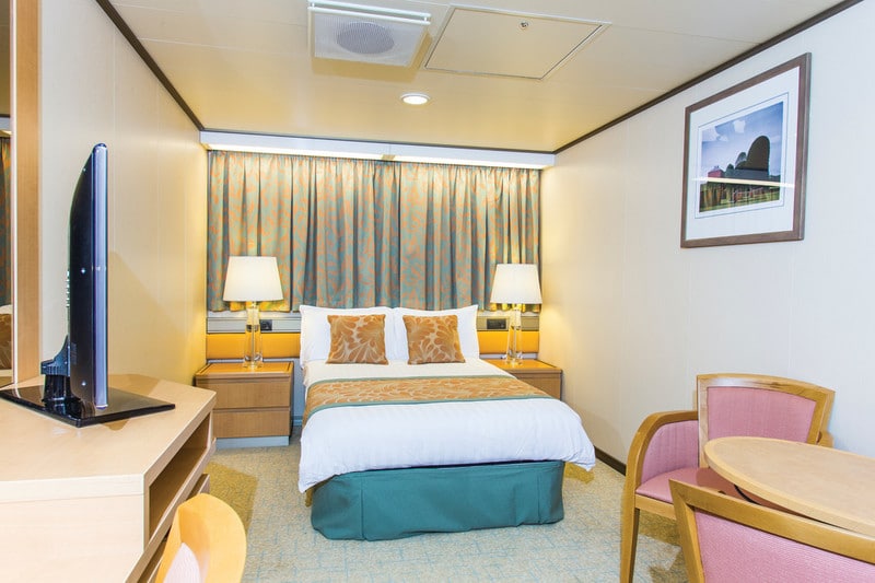 A Single cabin on the P&O Arcadia cruise ship, featuring a comfortable bed with orange and gold accent pillows and a matching throw. The room includes a small sitting area with a round table and two chairs, a flat-screen TV on a dresser, and bedside tables with lamps. The decor is warm and inviting, with patterned curtains and artwork adding a touch of elegance.
