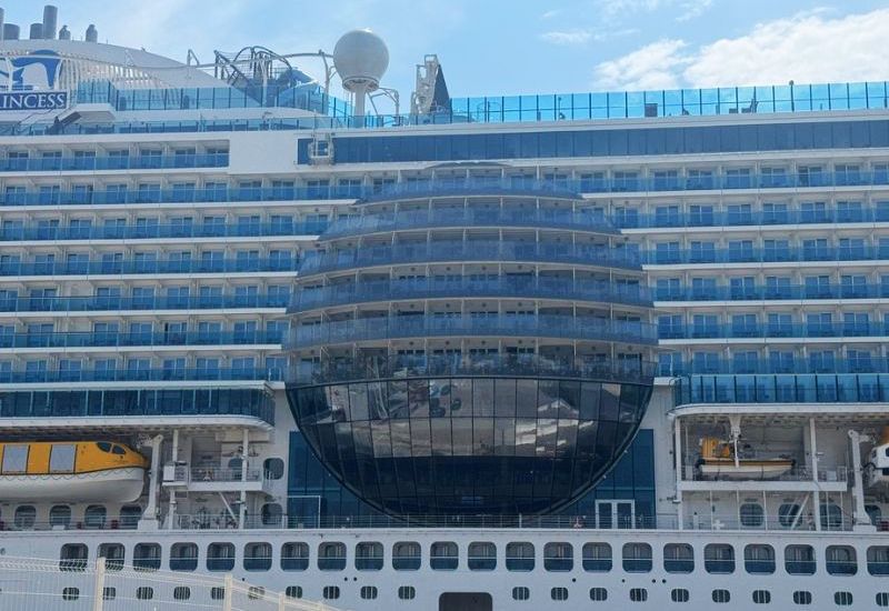The image shows a side view of Princess Cruises' Sun Princess, highlighting the unique "Sphere" structure at the center of the ship. This large, spherical feature is a prominent architectural element, with multiple decks of balconies curving around it. The balconies within this sphere offer guests panoramic views and a distinctive experience, likely enhancing the feeling of openness and connection with the ocean. Above and around the sphere, there are additional tiers of balconies, all part of the ship's accommodation layout. The sphere's reflective surface and modern design make it a standout feature on the ship, blending advanced engineering with luxury cruise amenities.