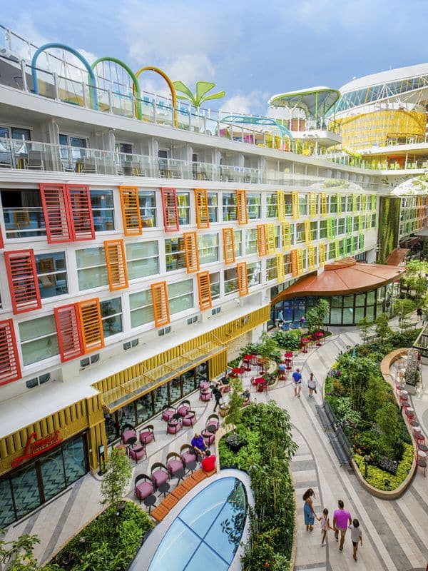 Lively central park area inside Royal Caribbean's 'Star of the Seas,' featuring multiple levels of staterooms with colorful shutters in red, orange, and yellow. The open-air atrium is bustling with activity, adorned with green plants, outdoor seating areas, and a curving pathway, creating a vibrant community space onboard the cruise ship.