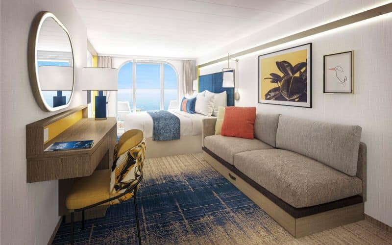 Balcony stateroom on Royal Caribbean's 'Icon of the Seas' featuring a stylish and spacious interior. This room offers a bed with blue and white bedding, a large gray sofa adorned with colorful throw pillows, and a balcony view of the ocean through floor-to-ceiling windows. The room is further enhanced by contemporary artwork, a wooden desk with a yellow chair, and a carpet with blue tones.