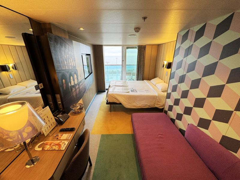 Interior of a balcony room on the Costa Smeralda cruise ship, featuring a large bed, geometric-patterned accent wall, a flat-screen TV, and a window with a view of the ocean.