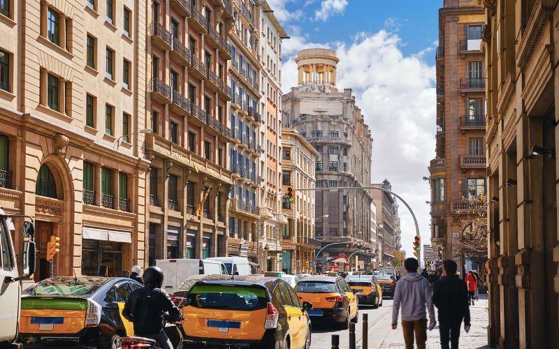 This image captures a bustling street scene in Barcelona, Spain, with iconic yellow-and-black taxis and pedestrians walking along a sunlit avenue. The street is lined with ornate historic buildings, and a prominent architectural landmark with a rounded tower is visible in the distance under a partly cloudy blue sky. The image conveys the vibrant urban atmosphere and charm of the city.
