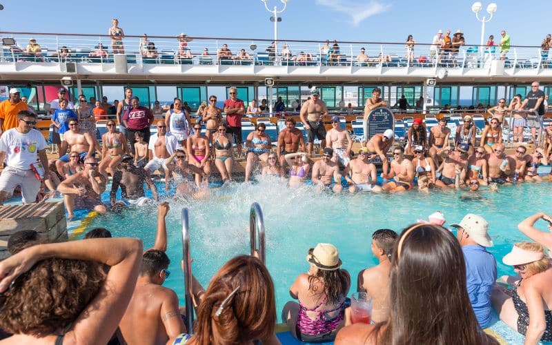 This image captures a lively scene at a cruise ship pool, with a large crowd of people gathered around and inside the pool. Many are sitting along the edge, dipping their feet in the water, while others are actively splashing and enjoying the sun. The atmosphere is festive and social, with people of all ages participating in the fun. The upper deck, visible in the background, is lined with additional passengers watching the action below, all under a bright, clear sky.