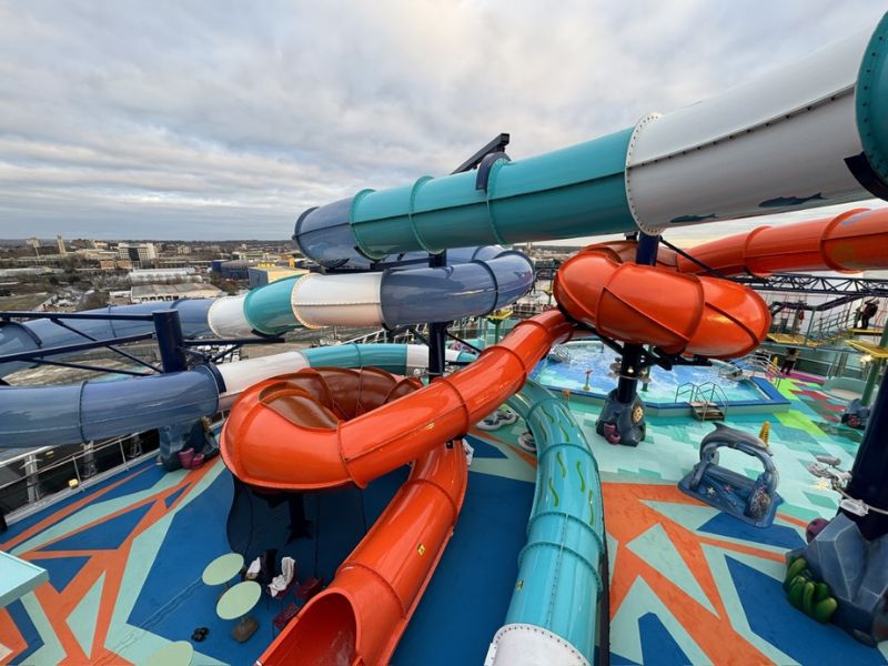 Alt text: "A dynamic view of a cruise ship's deck featuring a colorful array of winding waterslides in blue, teal, and orange. The slides spiral above a playful water park area, with a cityscape visible in the distance under a cloudy sky.