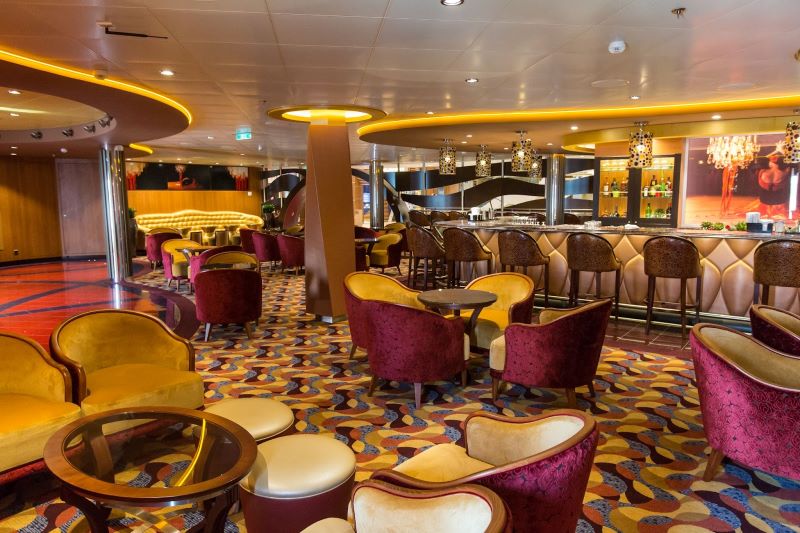 Inside the Boleros lounge on Royal Caribbean's Anthem of the Seas, featuring an eclectic mix of warm-toned velvet armchairs, a vibrant carpet with geometric patterns, and a sleek bar area illuminated by modern hanging lights, creating an inviting ambiance for socializing.