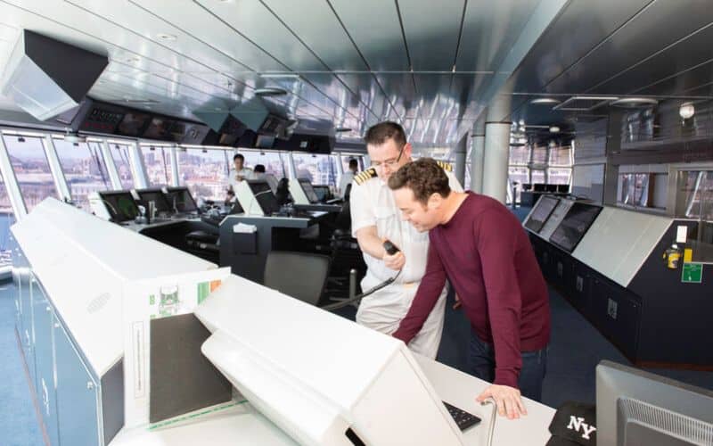 A ship captain and a passenger on the bridge of the P&O Britannia ship. The captain, dressed in a white uniform, is explaining the navigation controls to the passenger, who is leaning forward and engaged. The bridge features modern equipment and large windows offering a panoramic view.