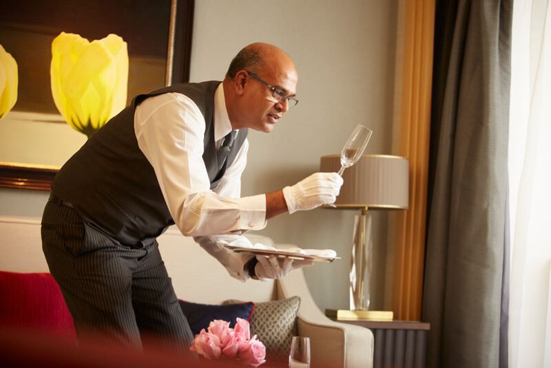 A professional butler on a Cunard cruise ship attentively inspects a wine glass for cleanliness. He is impeccably dressed in a vest and tie with white gloves, holding a tray with one hand, embodying the high standards of service and attention to detail that are hallmarks of luxury cruise hospitality.