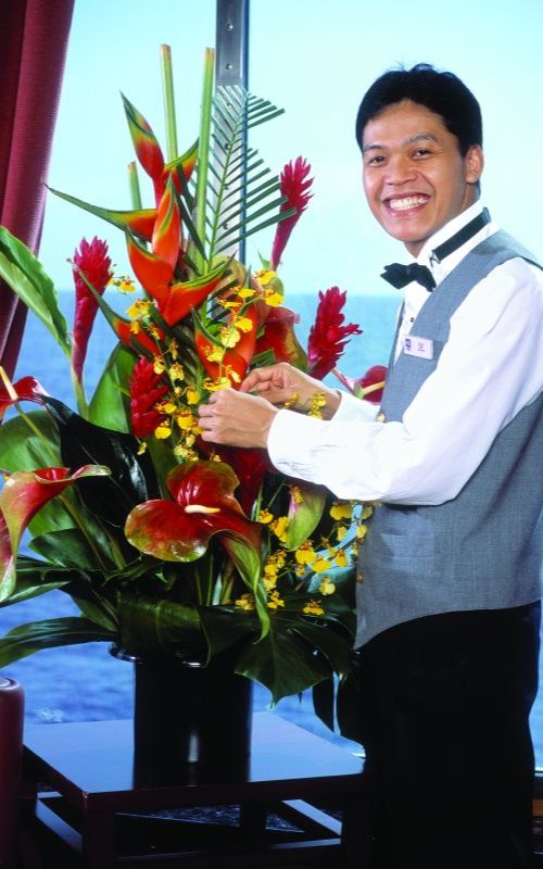 A smiling steward in a vest and bow tie arranges a vibrant bouquet of tropical flowers, including red ginger and yellow orchids, aboard an NCL cruise ship with a view of the sea through the window.