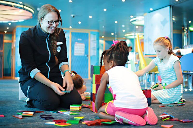 A Camp Ocean staff member on a cruise ship is sitting on the floor and engaging with a group of children who are building a structure with colorful blocks. The staff member is smiling and wearing a dark uniform with light blue trim, while the children, dressed in casual summer clothing, focus on their creative play. The background shows a bright and cheerful play area designed for kids' activities.
