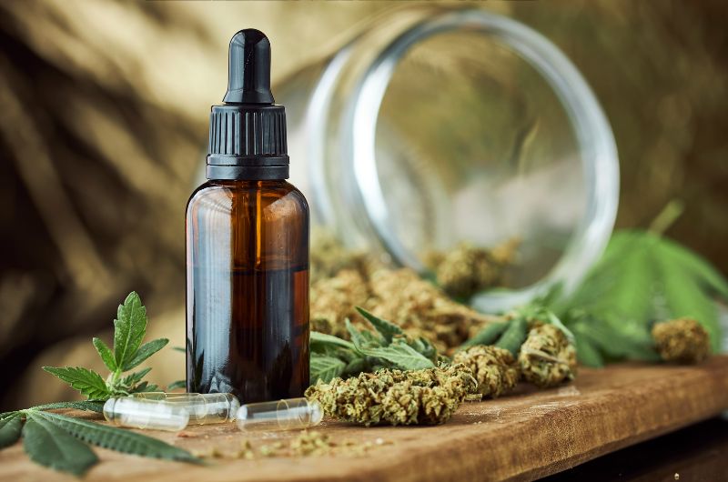 A bottle of CBD oil with a dropper cap is placed on a wooden surface surrounded by dried cannabis buds, leaves, and empty capsules. An open glass jar lies in the background, emphasizing the natural and holistic presentation of CBD products.