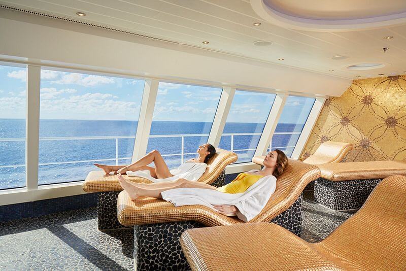 This image shows a serene spa area on a Carnival Cruise ship, featuring two women relaxing on heated mosaic-tiled loungers that overlook the ocean through large panoramic windows. The women are dressed in swimwear and white robes, enjoying the tranquil environment with a view of the open sea under a bright sky. The spa's interior has a luxurious feel, with soft lighting and elegant decor, providing a peaceful retreat for passengers.