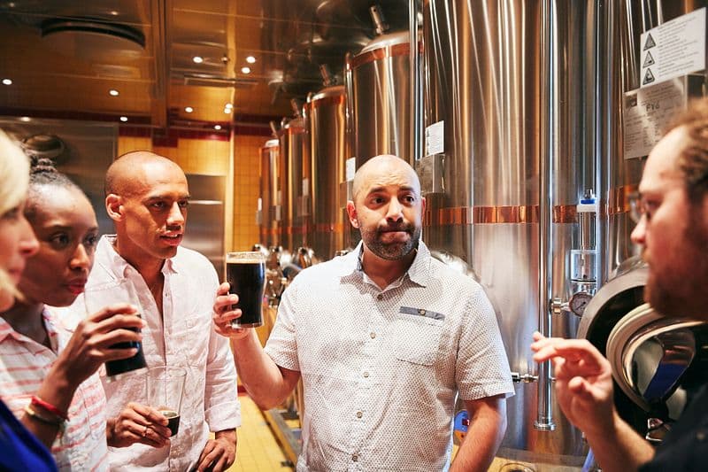 Guests engage in a beer tasting session at the RedFrog Pub and Brewery on Carnival Vista, surrounded by shiny brewing tanks, as an expert brewmaster in a patterned shirt explains the brewing process, enhancing the cruise experience with craft beer education.
