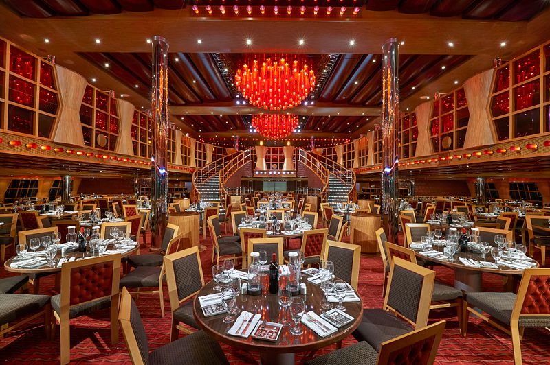 Elegantly set tables await guests in the main dining room of the Carnival Dream cruise ship, featuring rich wooden accents, a striking red and amber chandelier, and a grand double staircase, all creating a luxurious and inviting ambiance for a memorable dining experience.