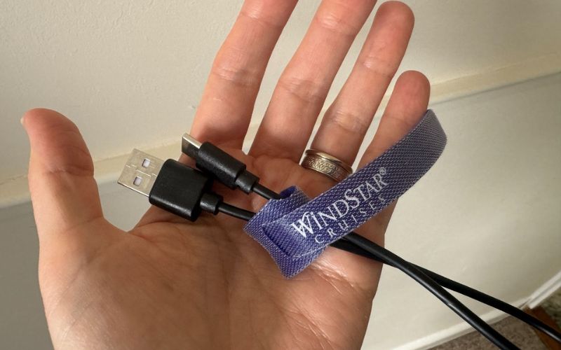 My open palm displays a multi-use charging cable with a USB on one end and both a USB-C and micro-USB connector on the other, neatly secured with a branded 'WINDSTAR CRUISES' cable tidy strap.