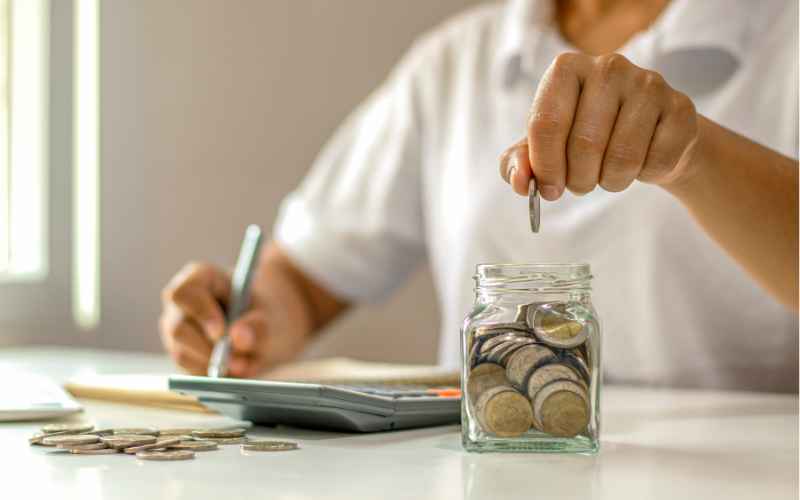 Someone putting some coins into a jar. They also are holding a pen and have a calculator.