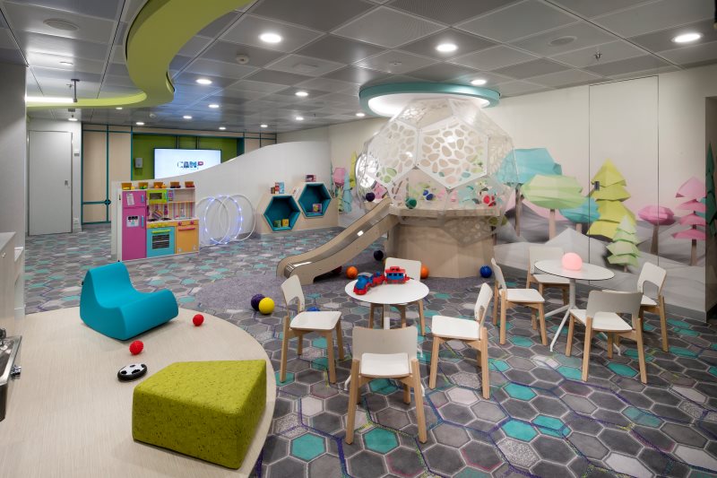 Camp at Sea on a Celebrity cruise ship, showcasing a playful and colorful indoor kids' area with geometric furniture, a whimsical slide and climbing structure, and interactive games for children to enjoy.