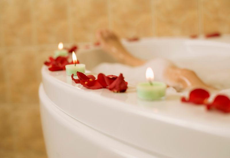 A relaxing scene with a bathtub filled with bubbles, adorned with lit green candles and scattered red rose petals around the edge. A person's legs are partially visible, enjoying a soothing bath in a serene and romantic atmosphere.