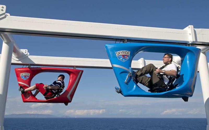 A passenger enjoys the SkyRide attraction on Carnival Panorama, pedaling a hanging bike along a track high above the ship's deck, offering expansive ocean views and a fun, aerial cycling adventure at sea.