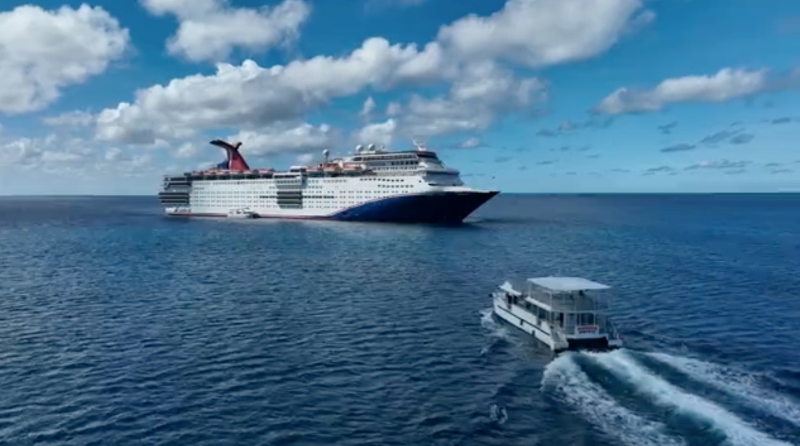 A Carnival Cruise ship with its distinctive red funnel at sea, with a smaller shuttle boat cruising swiftly in the foreground on the vast blue ocean under a partially cloudy sky.