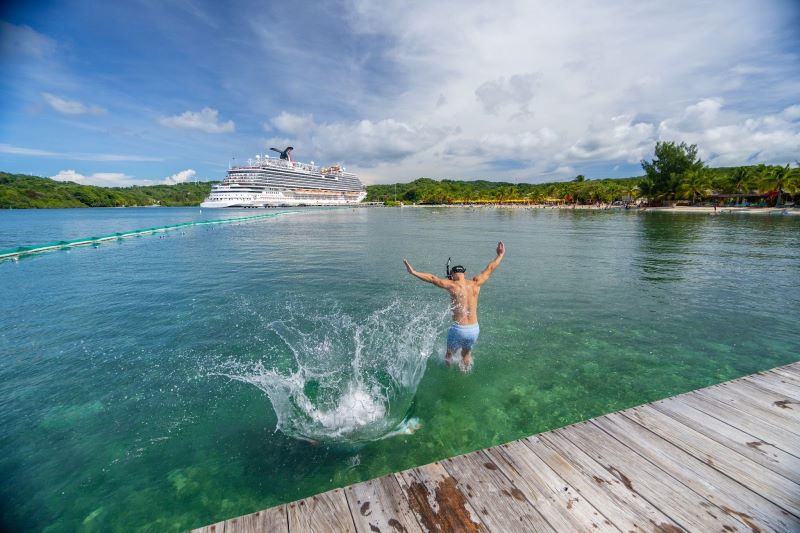 A person joyfully jumping into the clear tropical waters from a wooden pier, with a Carnival cruise ship anchored in the background near a lush, green coastline under a blue sky with scattered clouds.
