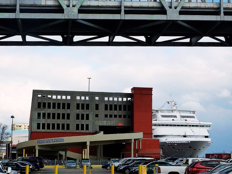 A Carnival cruise ship glimpsed beyond the Erato Street Cruise Terminal parking garage in New Orleans, showcasing the contrast between the bustling city infrastructure and the leisurely world of cruise travel.