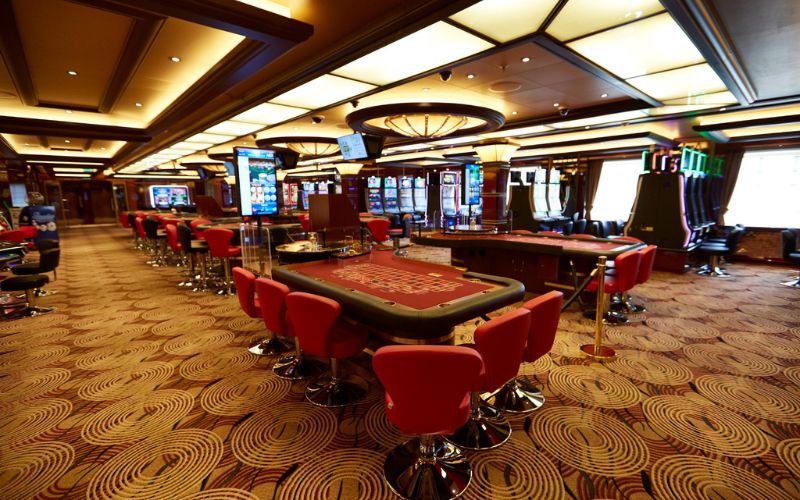 The vibrant casino on the Majestic Princess cruise ship, featuring red chairs at gaming tables, an array of colorful slot machines, and a luxurious, patterned carpet.