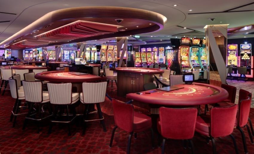 Inside the bustling casino on a Celebrity Cruise ship, the space is equipped with vibrant slot machines and gaming tables. The luxurious setting with red and cream decor invites guests for a night of entertainment and chance.
