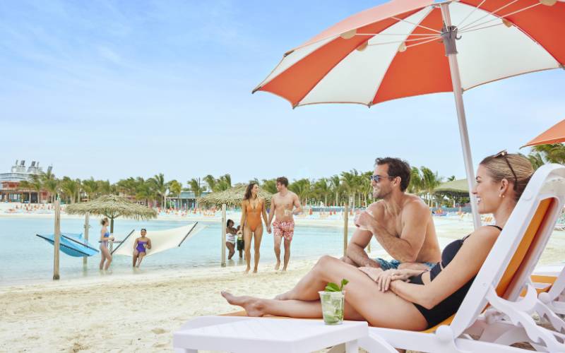 Cruise passengers are enjoying a sunny day at CoCo Cay beach. A couple relaxes on lounge chairs under a large orange and white umbrella, with a refreshing drink in hand. In the background, families and other beachgoers play in the clear blue water, walk along the sandy shore, and use hammocks strung between posts. The tropical setting includes palm trees and thatched umbrellas, creating a picturesque vacation scene.