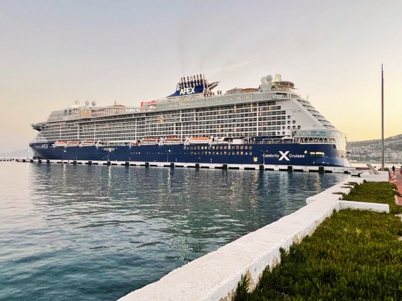The Celebrity Apex cruise ship from Celebrity Cruises moored at a quayside during dusk, its sleek design and illuminated windows standing out against the fading light, while the calm waters reflect its presence.