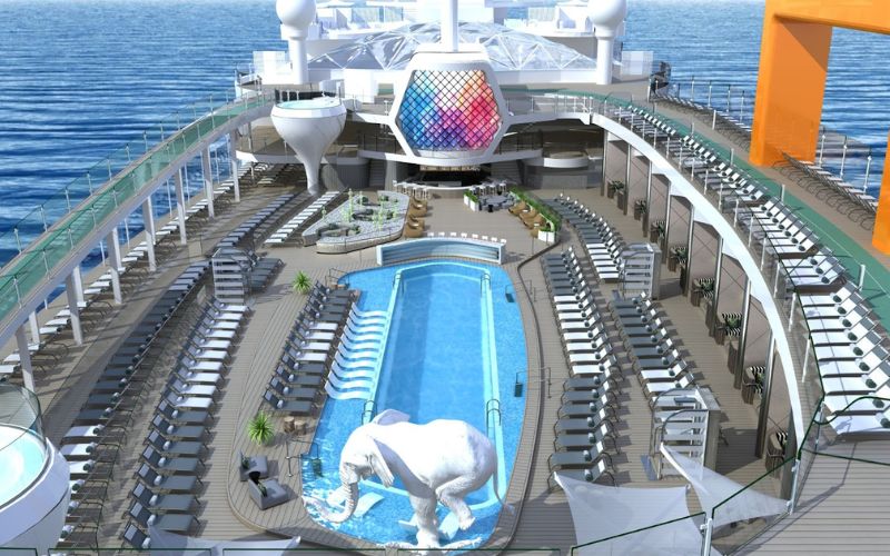 The pool deck of a Celebrity cruise ship, featuring an elegant oval pool with a striking white elephant sculpture, surrounded by neatly arranged sun loungers and shaded seating areas. Above, a distinctive geometric dome adds a modern touch to the ship's leisure amenities, all set against the tranquil ocean backdrop.
