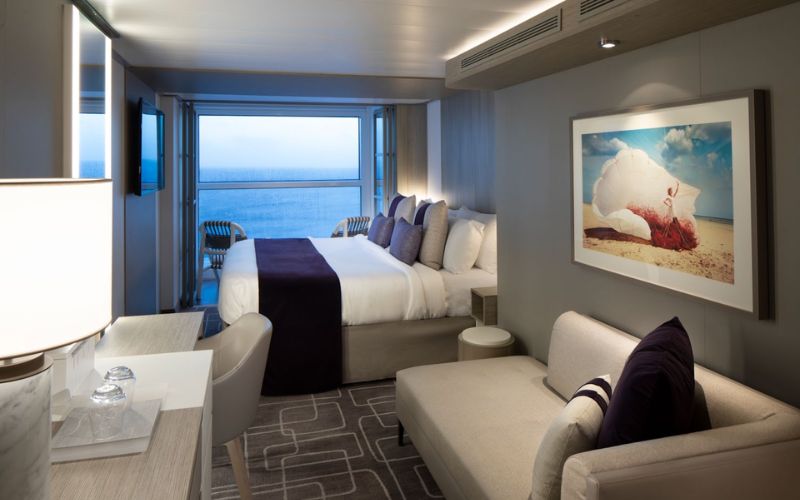 Interior of an Infinite Veranda stateroom on a cruise ship, featuring a large bed with white and navy bedding, a sofa, and a table set against the backdrop of a balcony overlooking the sea. The room is adorned with a framed picture of a seashell on the beach.