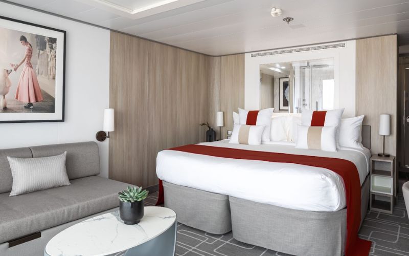 Modern and elegant cruise ship cabin with a red and white bed, a comfortable gray sofa, and a marble coffee table. A framed picture of a woman in a pink dress adds a touch of glamour, and the room's light wood tones create a warm and inviting atmosphere.