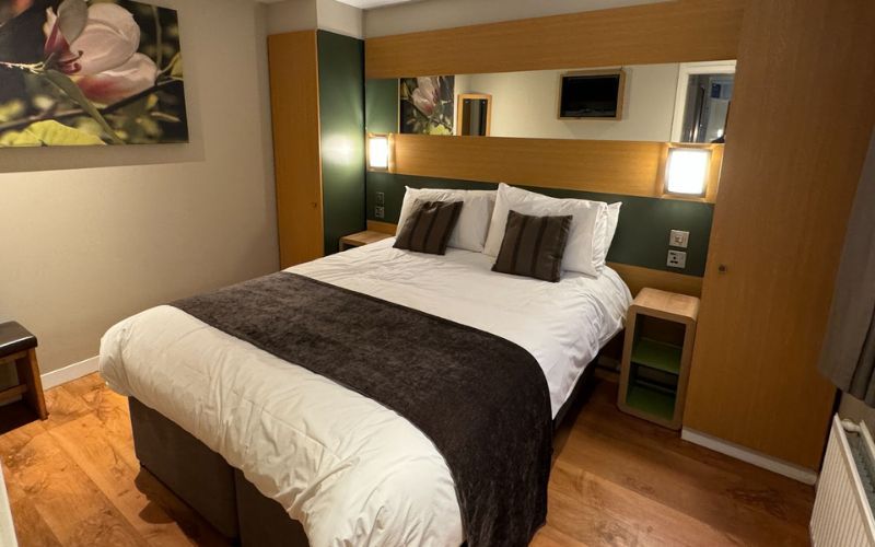 Modern bedroom featuring a large bed with white linens and a dark velvet throw, flanked by two bedside lamps. Above the headboard is a mirror, and the room is decorated with nature-inspired artwork, creating a serene and inviting atmosphere.
