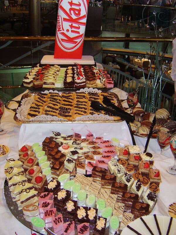 A lavish dessert buffet featuring a variety of intricately decorated cakes and pastries, with a prominent KitKat sign in the background. The assortment includes mini cheesecakes, chocolate slices, and colorful, frosted treats, arranged neatly on multiple tiers. The display is part of a chocoholics buffet on a P&O cruise ship.