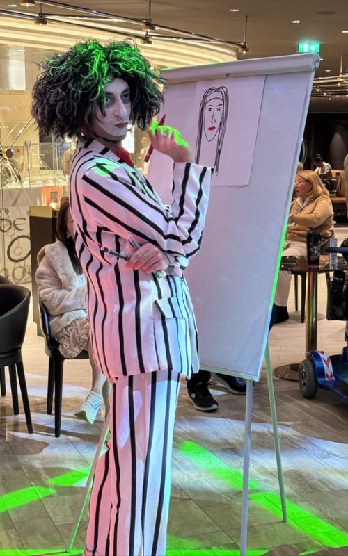 A performer in a black and white striped suit with a curly green wig poses beside an easel with a simple line drawing of a face, creating a whimsical and artistic atmosphere in a cruise ship's bustling indoor promenade